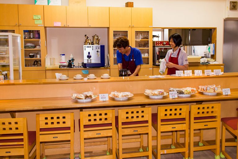 介護施設のカフェテリア。スタッフが飲料を準備し、カウンター前には様々なお菓子や食事が展示されている。